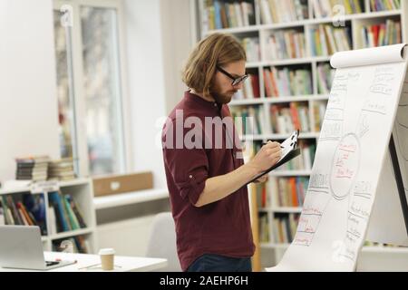 Young teacher in eyeglasses standing near the whiteboard and making notes in document he preparing for the lecture Stock Photo