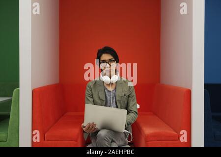 Portrait of Asian young businessman in headphones sitting on sofa and looking at camera while using laptop Stock Photo
