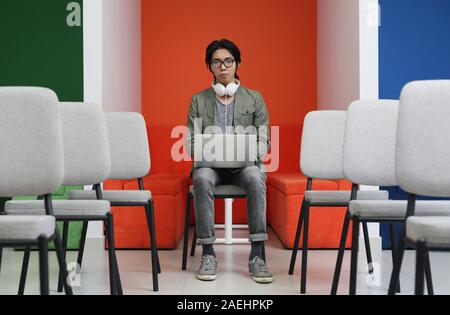 Portrait of serious Asian student in headphones sitting on chair and looking at camera while using his laptop computer Stock Photo
