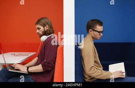 Young man sitting on sofa and reading a book with other young man sitting on sofa and using laptop they sitting back to back Stock Photo