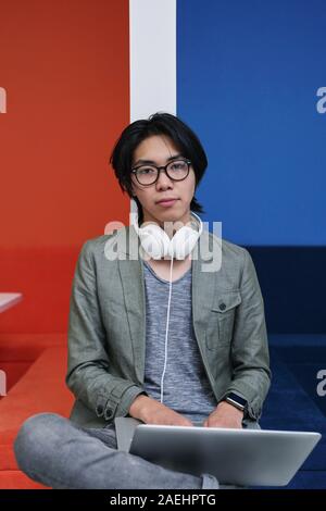 Portrait of Asian teenager in eyeglasses looking at camera while sitting on sofa and typing on laptop computer he studying online Stock Photo