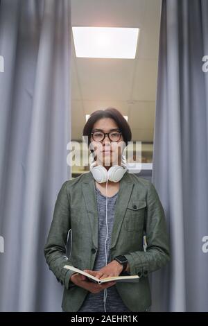 Portrait of Asian young man in headphones standing and looking at camera while reading a book Stock Photo
