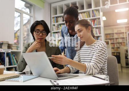 Young woman pointing at monitor of laptop and discussing new project together with her colleagues at the table in the library Stock Photo