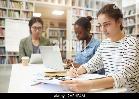Young woman sitting at the table and working with papers she preparing for exams together with her friends in the background Stock Photo