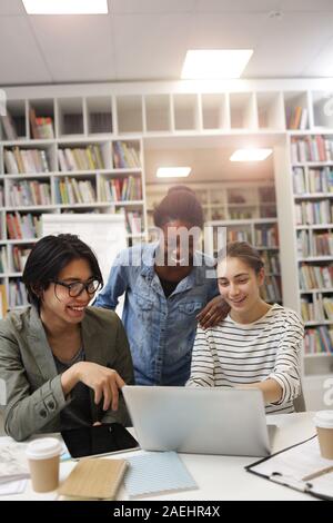 Group of young people laughing while watching online presentation on laptop at the table in the library Stock Photo