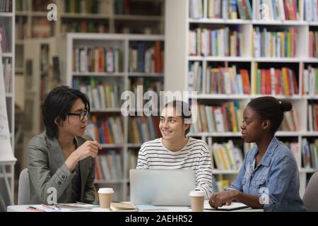Students sitting at the table discussing teamwork using laptop and drinking coffee in the library Stock Photo