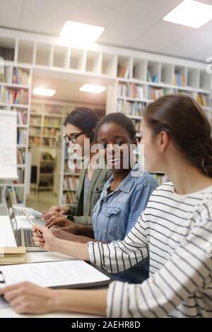 Two young women talking to each other and smiling while using laptop at the table with young man working in the background Stock Photo