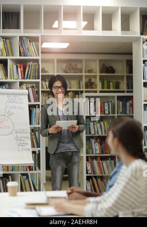 Asian smiling student standing near the whiteboard and presenting his report for other students in the library Stock Photo