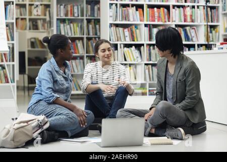Multiethnic group of students sitting on the floor using laptop and discussing their homework in team in the library Stock Photo