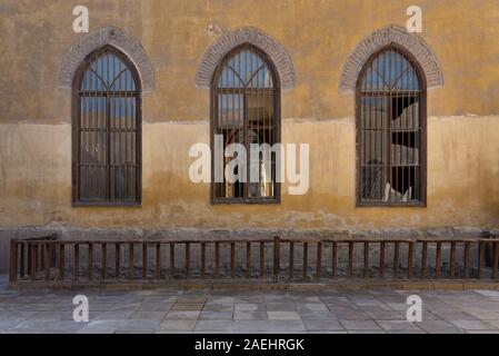 Row of three adjacent wooden grunge arched window with decorated wrought iron grid over yellow stone bricks wall and wooden balustrade, Medieval Cairo, Egypt Stock Photo