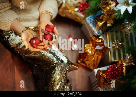 Closeup on stylish middle age housewife in gold sequin skirt and white sweater under decorated Christmas tree near present boxes holding broken Christ Stock Photo
