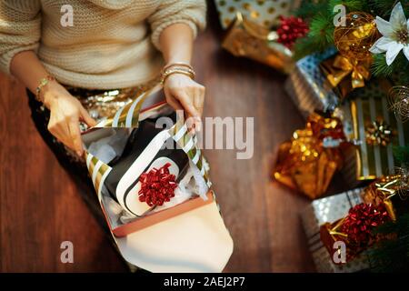 Closeup on trendy woman in gold sequin skirt and white sweater under decorated Christmas tree near present boxes wrapping Christmas gift VR for him. Stock Photo