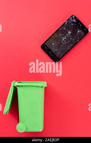 Broken screen mobile phone and green trash can on red background. Recycling. Stock Photo