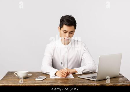Business, work and interview concept. Busy serious handsome, young asian man sitting desk in office drink coffee, writing report, studying documents Stock Photo