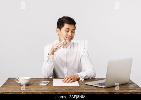 Pleased handsome young asian male entrepreneur, office worker sitting desk, smiling touched looking at cute kitten pictures via laptop, reading funny Stock Photo