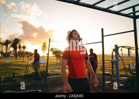 Sporty young man with earphones in his ears looking up at monkey bars for doing exercise in the gym park - Man motivating himself to do exercises Stock Photo