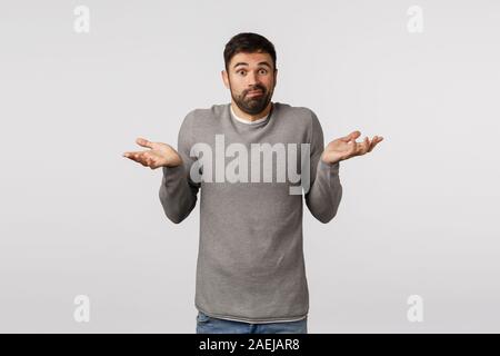 Guy asking help dont know what do. Perplexed, confused and indecisive cute clumsy bearded man in grey sweater, raise hands sideways, shrugging Stock Photo