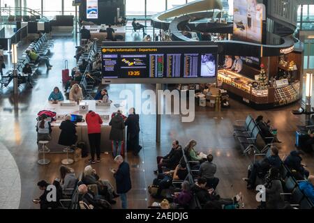 A flight information board in the middle of benches full of seated people waiting for their flights at London Heathrow Airport, Terminal 5. Stock Photo