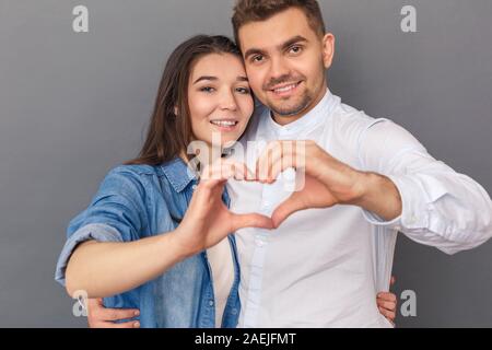 Relationship Concept. Young couple studio standing isolated on grey hugging making heart shape with hands smiling happy to camera close-up Stock Photo