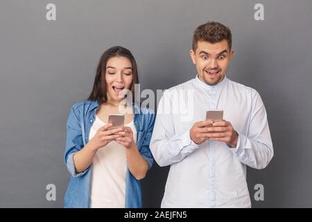 Using Devices. Young couple studio standing isolated on grey looking at screen of smartphone surprised promotion Stock Photo