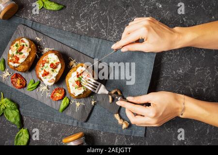 Baked stuffed potatoes with bacon, tomato and cheese on stone background. Female hands hold knife and fork. Top view Stock Photo