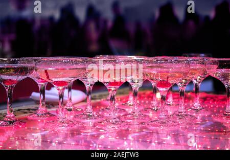 Many Champagne glasses at the party are lined up ready to be served. Celebrate, cheer, cocktail in the background. Elegant catering, tiered alcohol drinks on glass table in luxury purple concept. Stock Photo