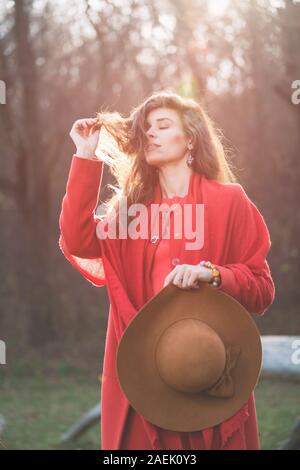 Fashion girl in red hold wide hat. Beautiful young brunette wears a red coat, scarf and brown wide-brimmed hat. Woman touches her hair, eyes closed. B Stock Photo