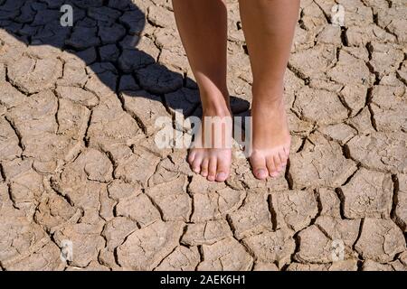 Close up photo of a woman’s feet standing barefoot on a cracked earth, dried out land. Lack of rainfall takes its toll, causing desertification to the Stock Photo