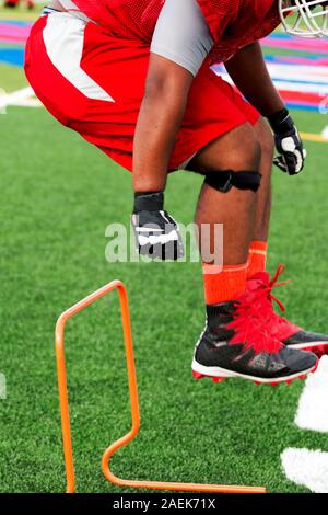 A high school football player is cross training by jumping over orange mini hurdles on a green turf field during practice. Stock Photo