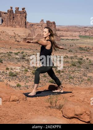 Young woman practices yoga at the La Sal Mountains Overlook in Arches National Park near Moab, Utah. In the background are the Courthouse Towers. Stock Photo