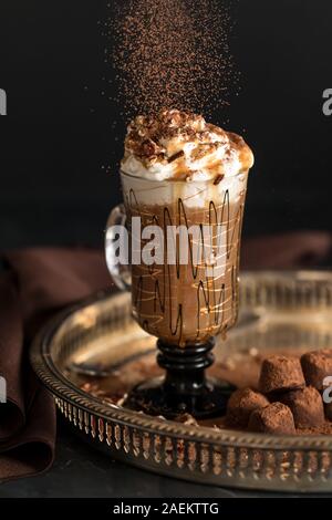 A front facing close up of a decorative glass mug filled with hot cappuccino and topped with whip cream, crushed pecans and cocoa powder being poured Stock Photo