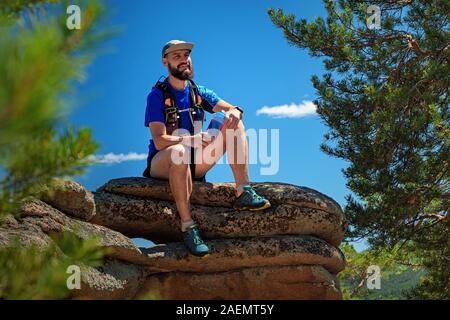 A male runner is sitting on the edge of a cliff, he is resting after a successful workout. Outdoor trail running. Man in blue t-shirt and black shorts Stock Photo
