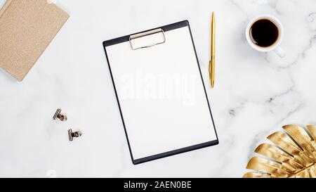 White marble desk table with blank paper clipboard mockup, golden tropical monstera leaf, cup of coffee and office supplies. Beauty blogger workspace, Stock Photo