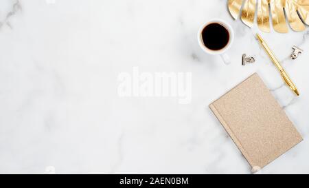 White marble desktop with golden office accessories and stationery. Home office desk table with gold tropical monstera leaf, cup of coffee, paper note Stock Photo