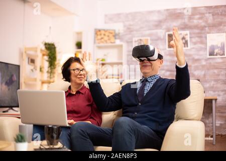 Old elderly retired man using VR virtual reality headset in their cozy apartment. Stock Photo