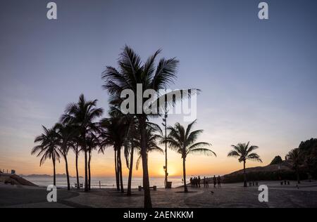 Sunrise on the Arpoador boulevard with the Devils beach and silhouetted palm trees in Rio de Janeiro against a clear orange and blue sky