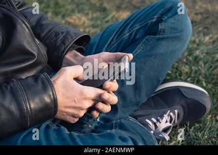 Caucasian man wearing a leather jacket and blue jeans, sitting on the grass, using a mobile phone in a park. Communication in a natural environment. Stock Photo