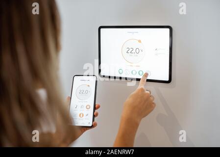 Young woman controlling temperature in the living room with smart phone and digital touch screen panel. Concept of heating control in a smart home Stock Photo