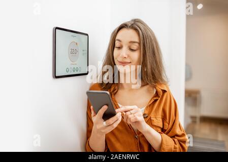 Young woman controlling temperature in the living room with smart phone and digital touch screen panel. Concept of heating control in a smart home Stock Photo
