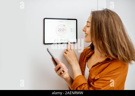 Young woman controlling temperature in the living room with smart phone and digital touch screen panel. Concept of heating control in a smart home Stock Photo