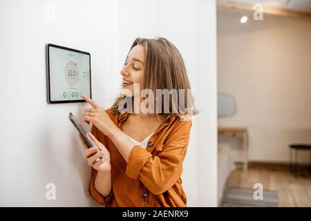 Young woman controlling temperature in the living room with smart phone and digital touch screen panel. Concept of heating control in a smart home Stock Photo