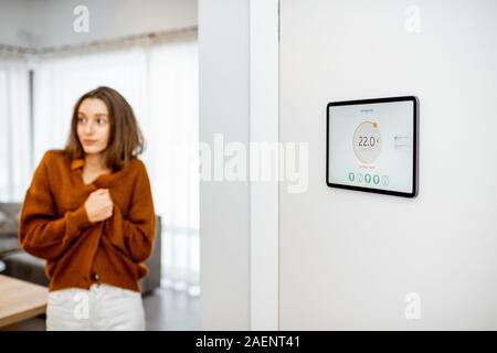 Digital panel for smart home managing with launched heating control application in the living room. Woman feeling cold on the background Stock Photo