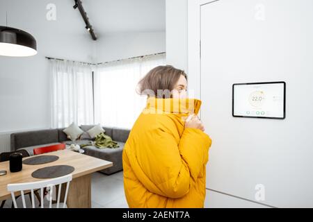 Young woman in winter clothes, feeling cold, waiting for the house to heat up, controlling heating system with a smart home devices Stock Photo