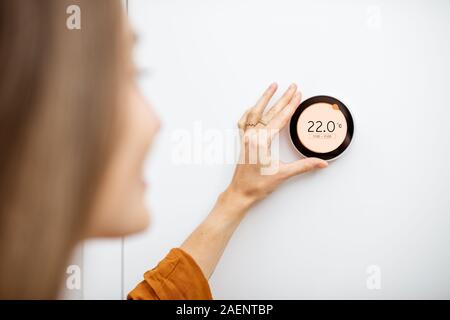 Young woman regulating heating temperature with a modern wireless thermostat installed on the white wall at home. Smart home heating regulation concept Stock Photo
