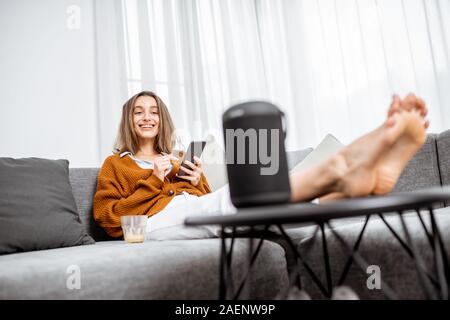 Young cheerful woman controlling home devices with a voice commands, talking to a smart column while relax on the couch at home Stock Photo