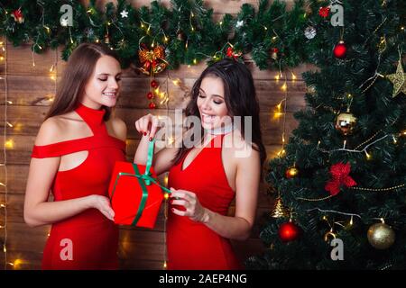 Pretty young woman giving an attractive young friend a surprise gift gift-wrapped with red paper near christmas tree, Stock Photo
