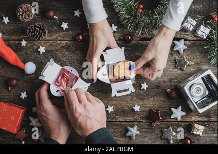 Top view of male and female hands packing home made cookies and candy canes in small gift boxes on rustic wooden desk with Christmas decoration and or Stock Photo