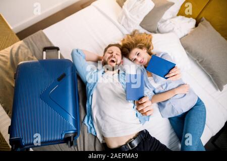 A happy young couple on their vacations lying on the bed in hotel room and holding blue passports. Focus on passports in hands. Stock Photo