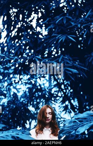 Beautiful young redhead woman in tropical garden toned in blue. Stock Photo