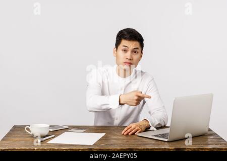Serious displeased young asian businessman sitting office desk, pointing laptop screen and looking strict camera, frowning skeptical, scolding Stock Photo